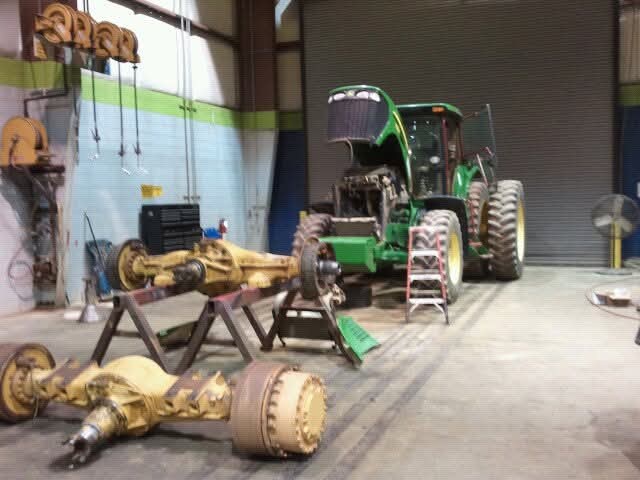 A green tractor with its hood open and two heavy axles on stands in a shop bay